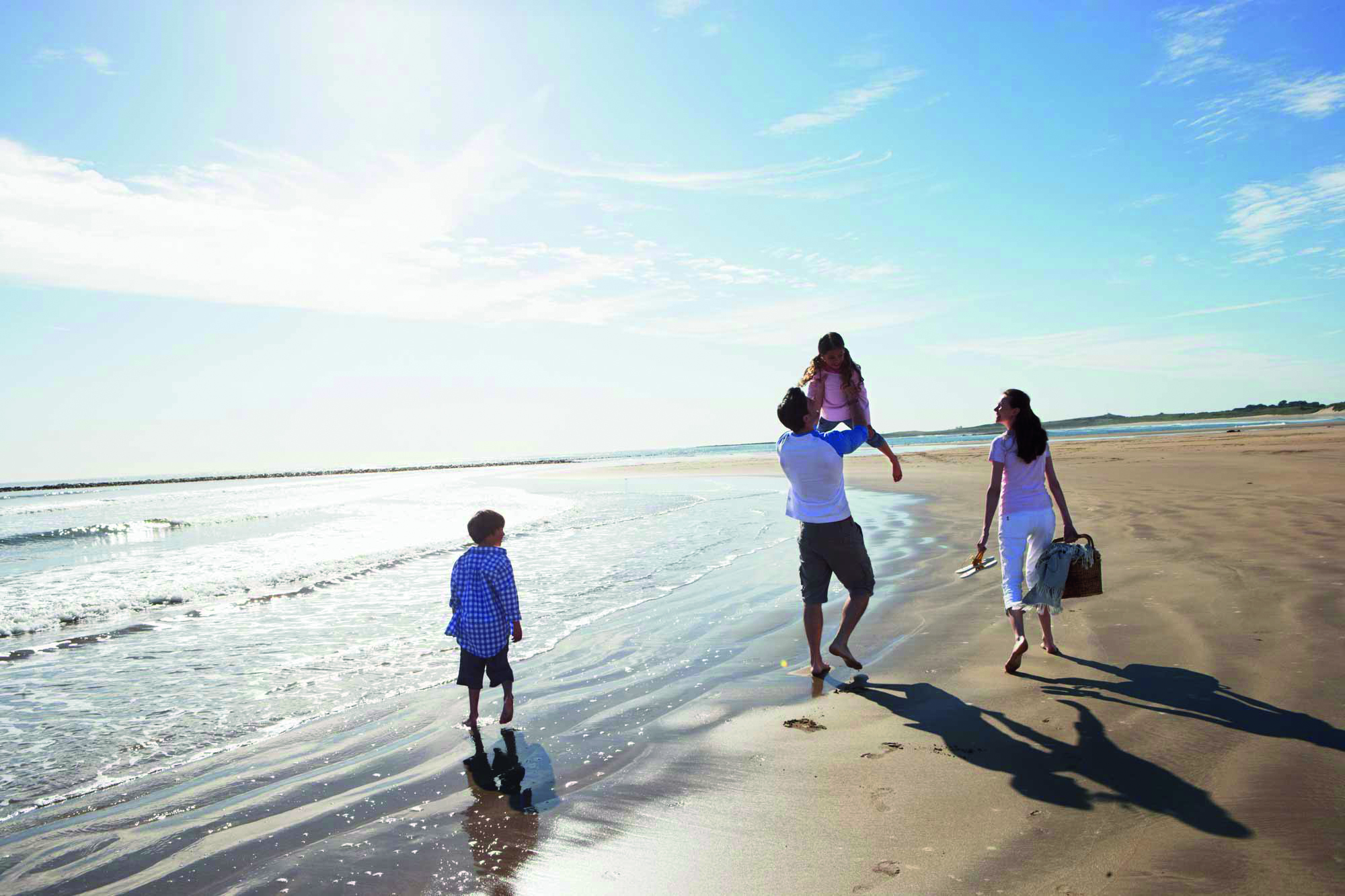 Rear View Of Family Walking Along Beach With Picnic Basket